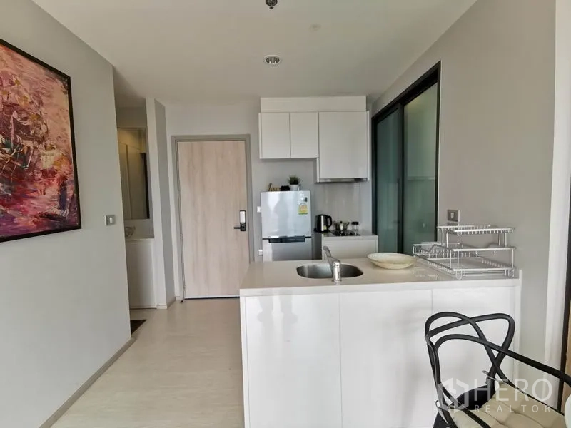 Condominium for rent in Khlong Toei, Bangkok - Compact white kitchen with island sink, upper cabinets and refrigerator.