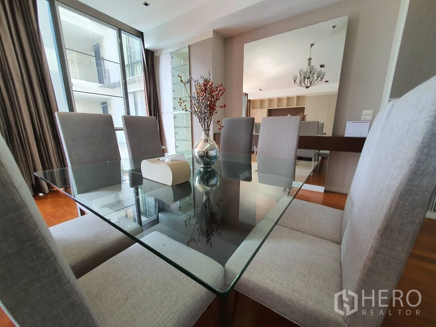 Condominium for rent in Khlong Toei, Bangkok - Dining area with a glass-top table, six upholstered chairs and a large mirror beside full-height windows.