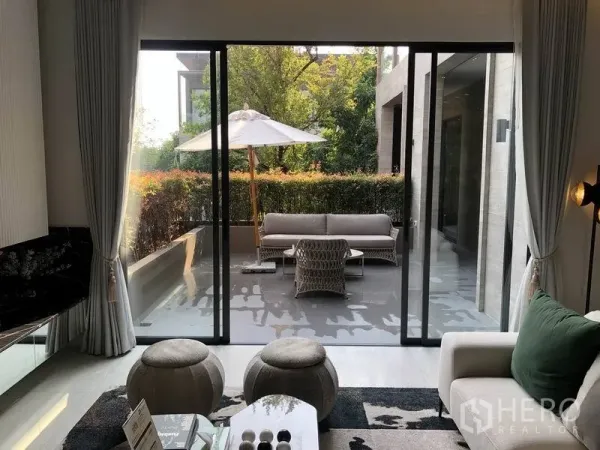 独立式住宅 出售在Bang Kapi, Bangkok - 客厅连接滑动玻璃门，外有遮阳伞、户外沙发与绿篱庭院。