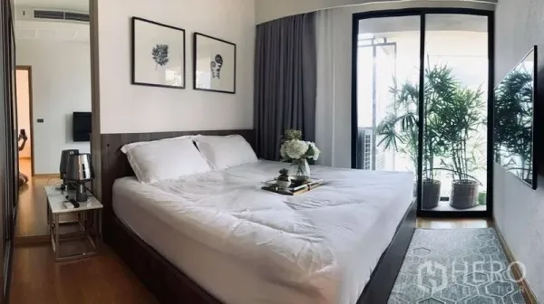 Condominium for rent in Watthana, Bangkok - Bright master bedroom with king bed, wall art and sliding doors to a balcony lined with potted plants.