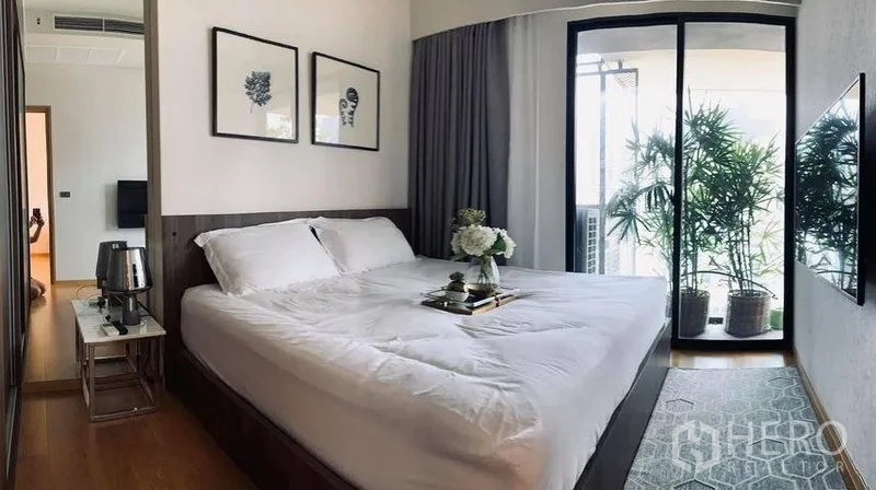 Condominium for rent in Watthana, Bangkok - Bright master bedroom with king bed, wall art and sliding doors to a balcony lined with potted plants.