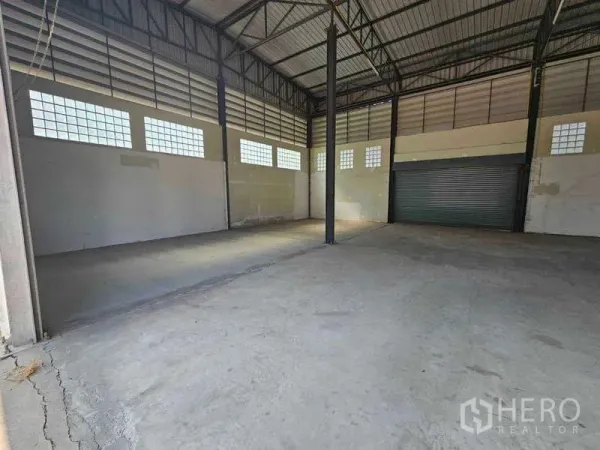 Warehouse for rent in Bang Bua Thong, Nonthaburi - High-clearance warehouse interior with steel truss roof, concrete floor, glass-block windows and a roller shutter.