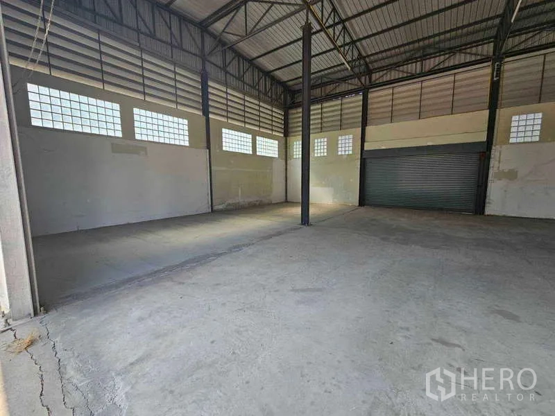 Warehouse for rent in Bang Bua Thong, Nonthaburi - High-clearance warehouse interior with steel truss roof, concrete floor, glass-block windows and a roller shutter.