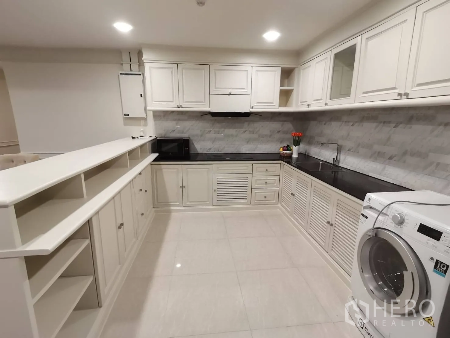 Condominium for rent in Watthana, Bangkok - Spacious kitchen with white cabinetry, black countertops, tiled backsplash, open shelving, and a washing machine.