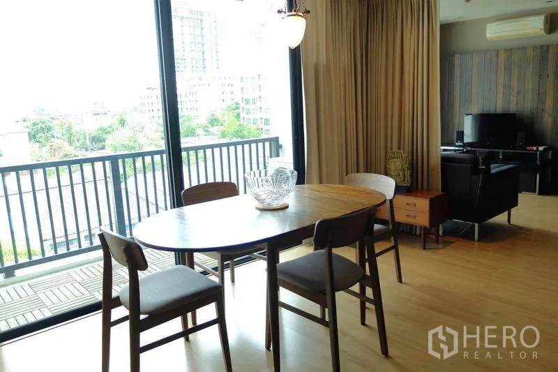 Condominium for sale in Watthana, Bangkok - Dining table by floor-to-ceiling windows opening to a balcony with city and greenery views.
