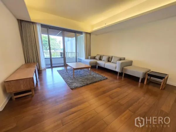 Condominium for rent in Yan Nawa, Bangkok - Bright living room with L-shaped sofa, wood floors and floor-to-ceiling sliding doors opening to a balcony with garden views.