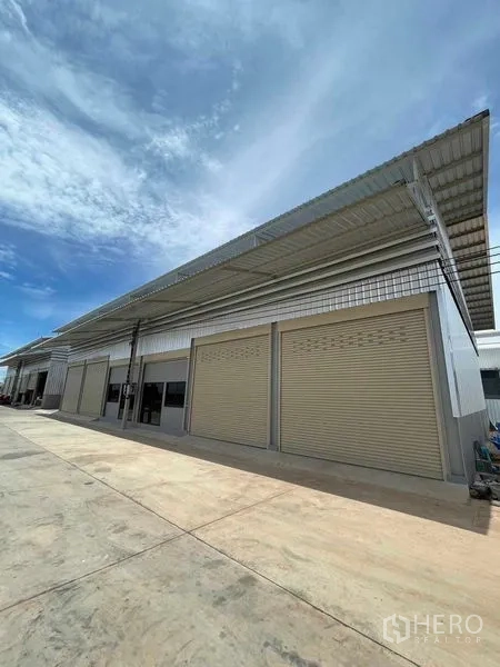 Warehouse for rent in Phra Samut Chedi, Samut Prakan - Exterior of a newly built industrial warehouse with multiple electric roller shutters and a wide concrete driveway under a blue sky.