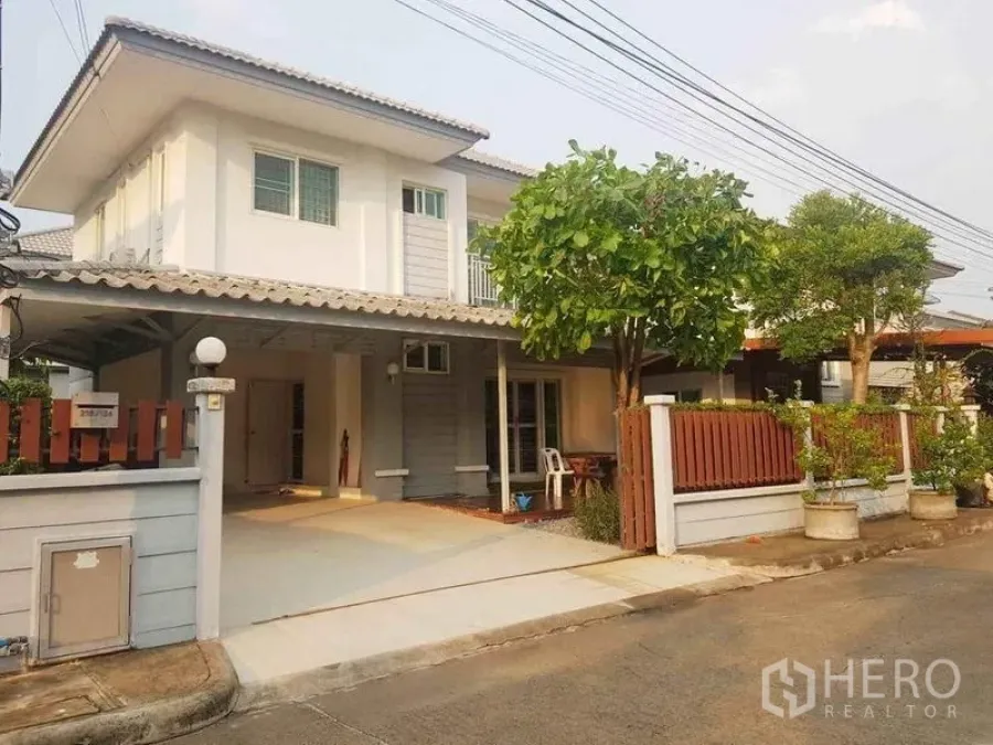 Detached House for sale in Nonthaburi - Front view of the house with wide driveway, covered carport, trees and fence.