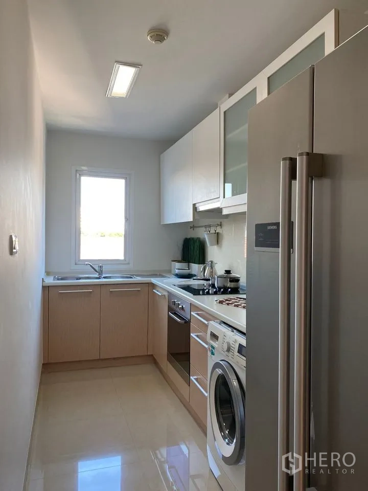 Condominium for rent or sale in Phra Khanong, Bangkok - Windowed galley kitchen with light wood cabinets, oven, cooktop, washing machine and double-door fridge.