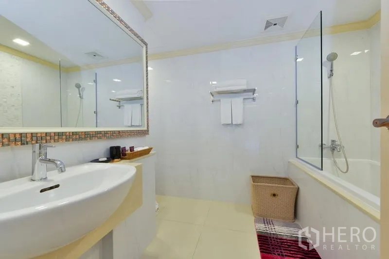 房间 出租在Khlong Toei, Bangkok - 浴室盥洗台，宽大镜子、收纳篮与玻璃浴缸淋浴区。