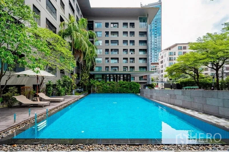 คอนโด ขาย - สระว่ายน้ำกลางคอนโด รายล้อมด้วยต้นไม้ เตียงอาบแดด และอาคารสูงกลาง
