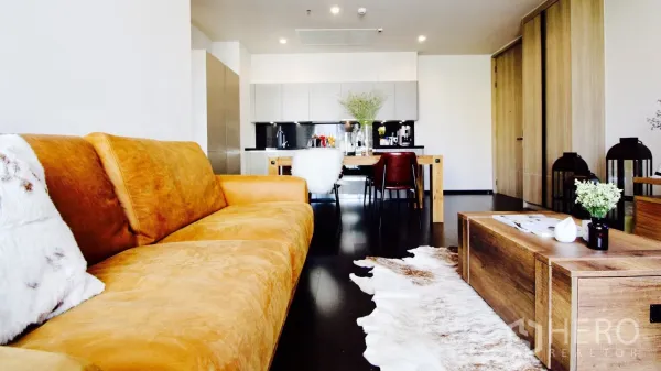 Condominium for rent or sale in Watthana, Bangkok - Spacious living room with a tan leather sofa, wood coffee table, and modern kitchen in the background.