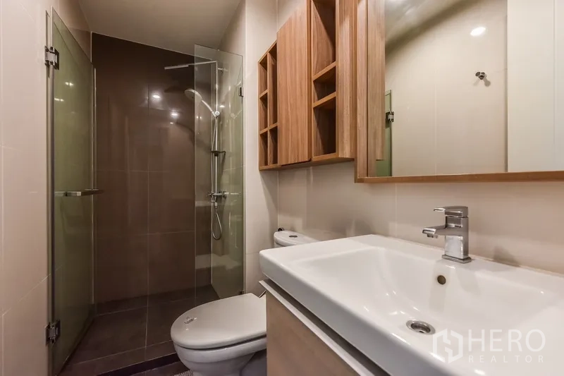 Condominium for rent or sale in Huai Khwang, Bangkok - Modern bathroom with glass-enclosed shower, vanity sink, and wood storage cabinets.