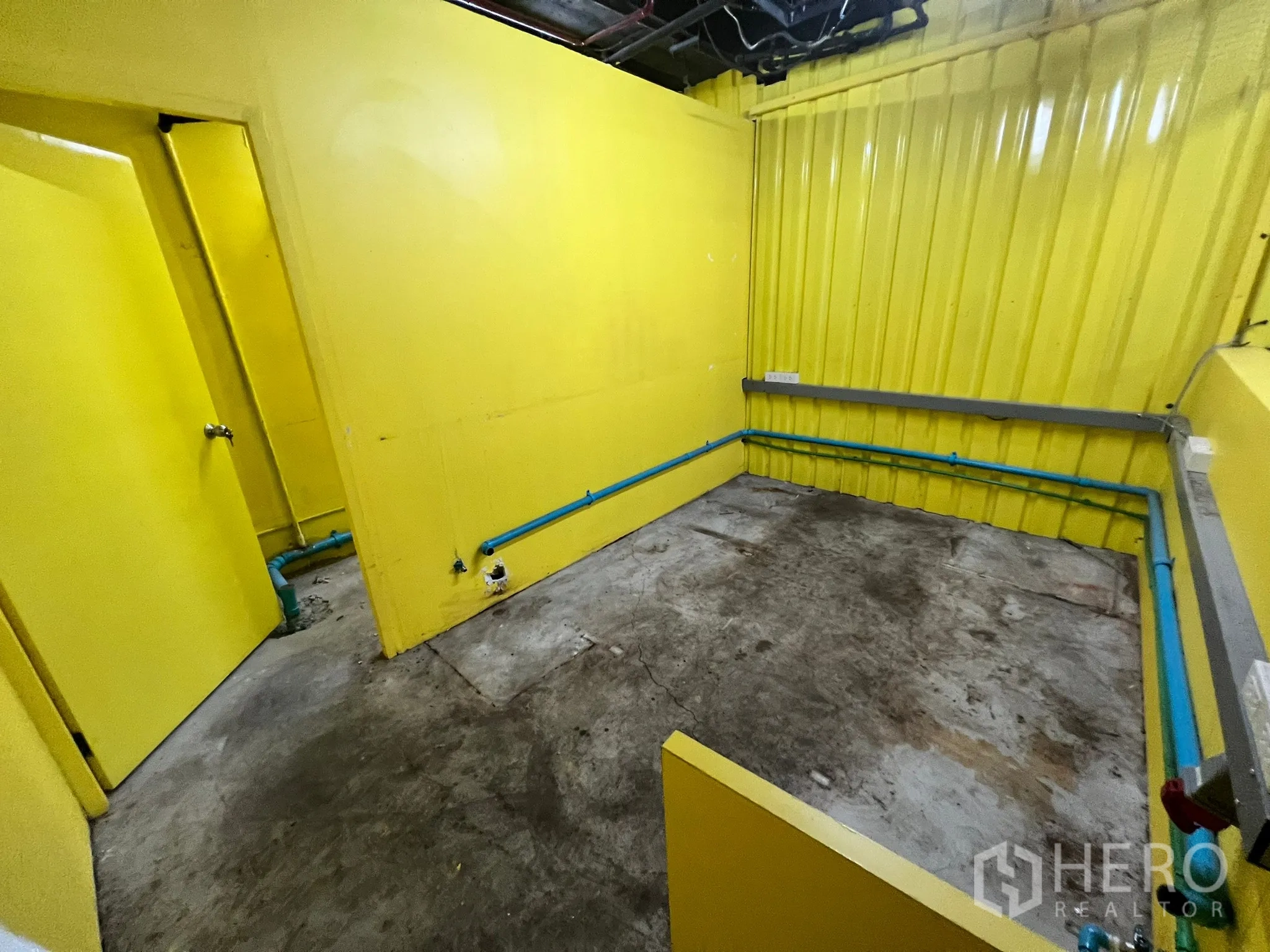 พื้นที่ขายปลีก ให้เช่า - ห้องยูทิลิตี้ด้านหลัง ผนังสีเหลืองสด ท่อประปาสีฟ้า และพื้นคอนกรีต พร้อมปรับแต่ง