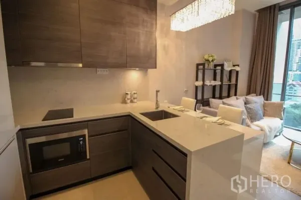 Condominium for sale in Watthana, Bangkok - Modern U-shaped kitchen with quartz countertops, built-in microwave, induction hob and breakfast bar opening to a bright living area with balcony view.