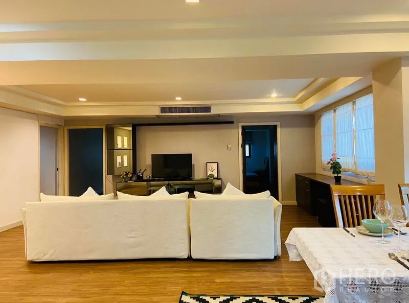 Condominium for rent in Watthana, Bangkok - Living room facing a TV wall unit, wide windows with Roman shades, and a white sofa on timber flooring.