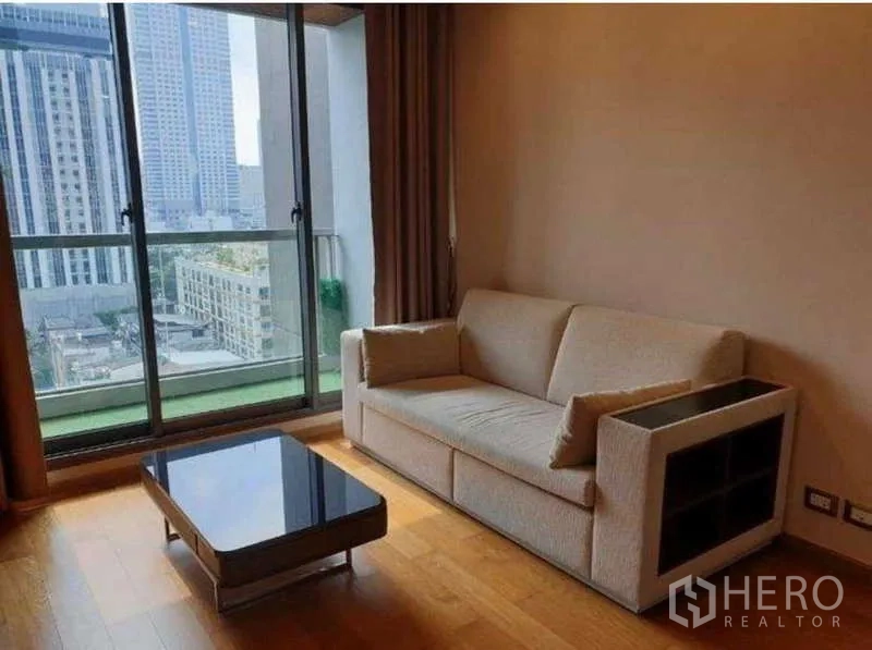 Condominium for rent in Bang Rak, Bangkok - Bright living room with beige sofa, glass coffee table and sliding doors to a balcony with Sathorn city skyline.
