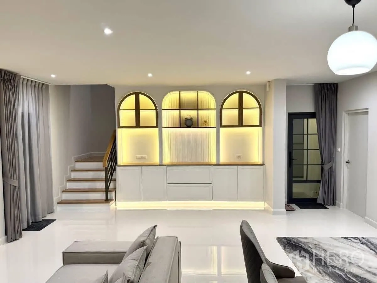 Detached House for rent in Bang Phli, Samut Prakan - Bright living–dining wall with arched backlit display cabinets and adjacent staircase.