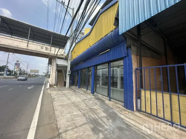 货栈 出租在Pak Kret, Nonthaburi - 临街仓库外观，黄色与蓝色立面，整面玻璃展示窗，旁有人行天桥。