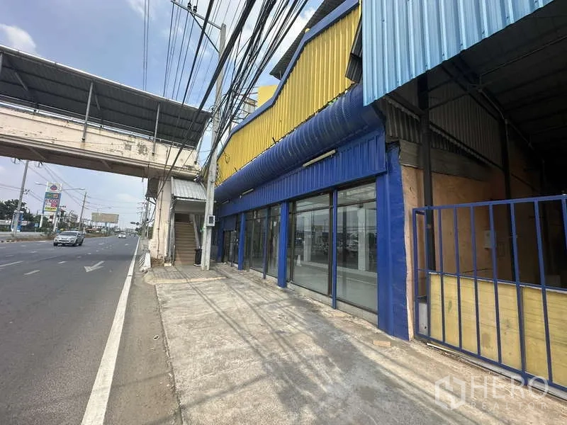 Warehouse for rent in Pak Kret, Nonthaburi - Street-facing warehouse exterior with blue and yellow facade and full-height glass frontage beside a pedestrian bridge.