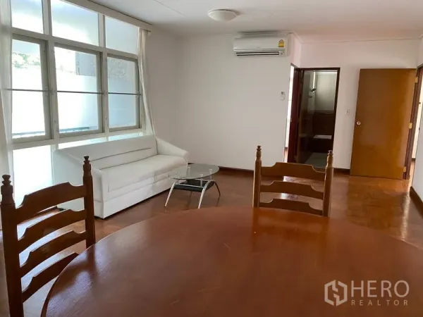 Apartment for rent in Khlong Toei, Bangkok - Open-plan living viewed from dining table, large windows with curtains and wall-mounted air conditioner.
