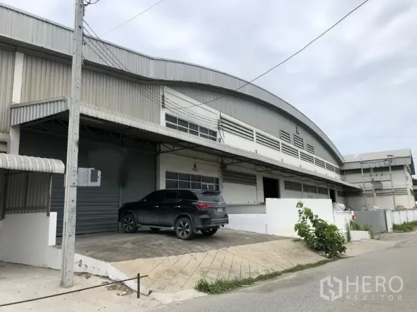 Warehouse for rent in Bang Bo, Samut Prakan - Front facade of the warehouse with curved metal roof, roller shutters and a small parking ramp.