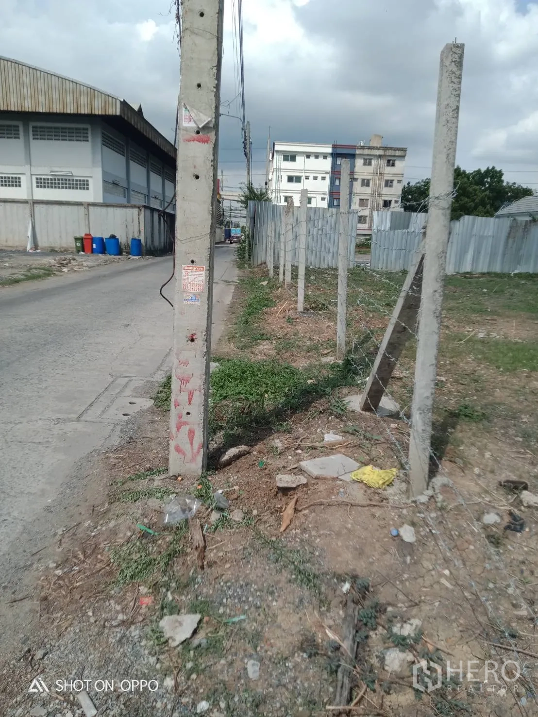 Land for rent in Khan Na Yao, Bangkok - Corner of the plot at a utility pole with barbed‑wire posts and a warehouse on the left.