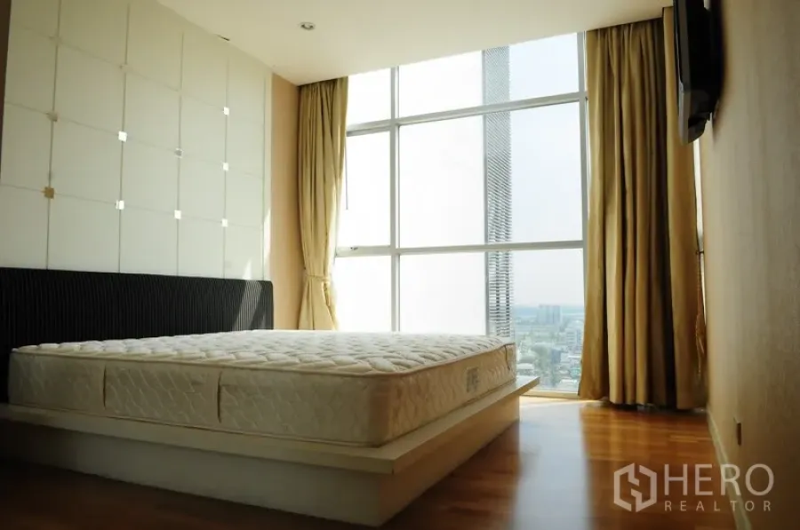 Condominium for rent in Pathum Wan, Bangkok - Sunlit bedroom with floor-to-ceiling windows, city view, curtains and warm wood flooring.