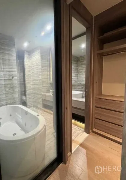 Condominium for rent in Watthana, Bangkok - Walk-in closet leading to a spa-style bathroom with a jetted bathtub behind a glass partition and wood cabinetry.