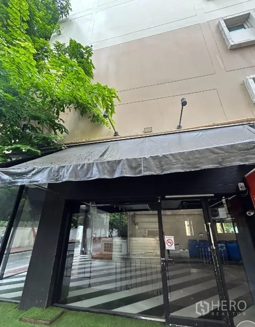 零售空间 出租在Bang Rak, Bangkok - 玻璃入口特写，深色遮阳篷与树荫覆盖。