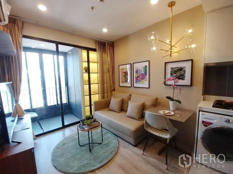 Condominium for rent in Huai Khwang, Bangkok - Cozy lounge with sofa and dining set, modern chandelier, kitchenette with washing machine, and sliding doors to the balcony.