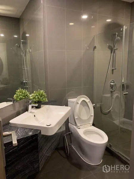 公寓 出租在Bang Rak, Bangkok - 现代浴室，玻璃淋浴间、挂墙式台盆与灰色瓷砖。