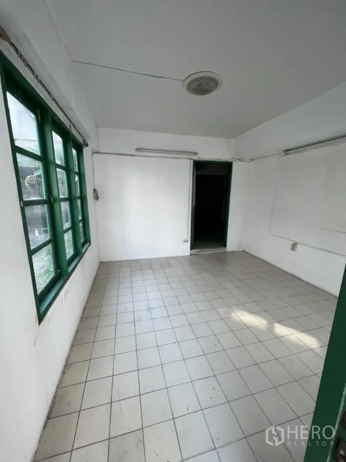 Townhouse for sale in Watthana, Bangkok - Empty room with white walls, tiled floor, and green-framed windows ready to refurbish.