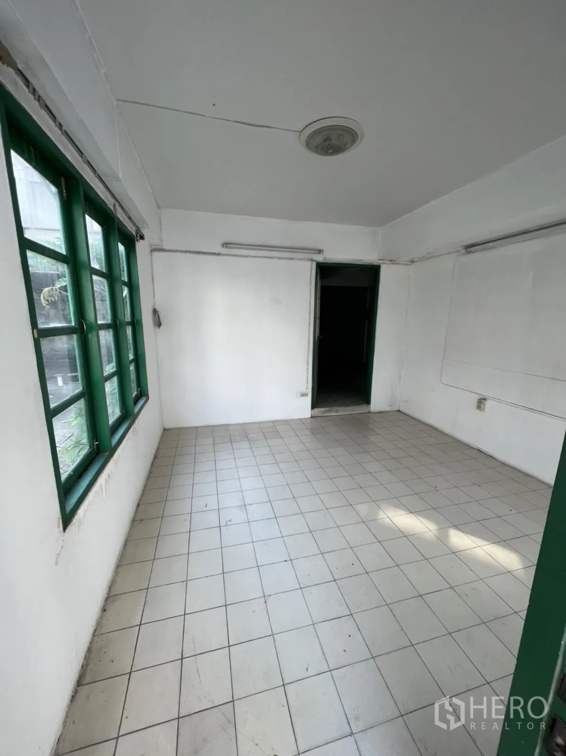 Townhouse for sale in Watthana, Bangkok - Empty room with white walls, tiled floor, and green-framed windows ready to refurbish.