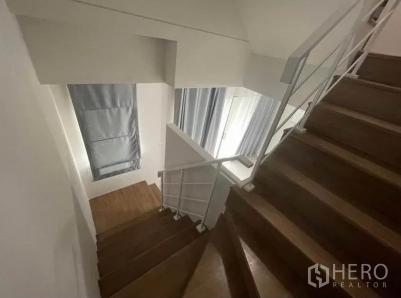 Townhouse for rent in Phasi Charoen, Bangkok - Wooden staircase with white metal railing and natural light from tall windows.