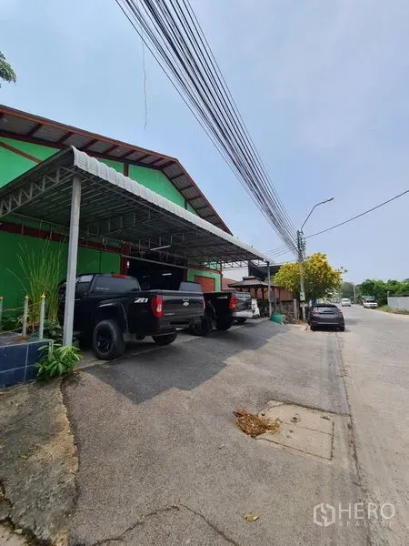 Warehouse for sale in Mueang Samut Prakan, Samut Prakan - Street-side view of a green warehouse with metal canopy parking and pickup trucks along a quiet road in Pak Nam, Samut Prakan.