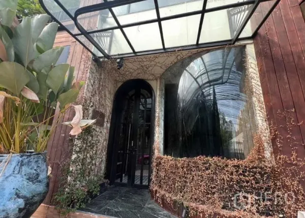 零售空间 出租在Watthana, Bangkok - 店铺入口设拱形黑门与弧形橱窗，配玻璃雨棚、攀缘植物和大型花盆。