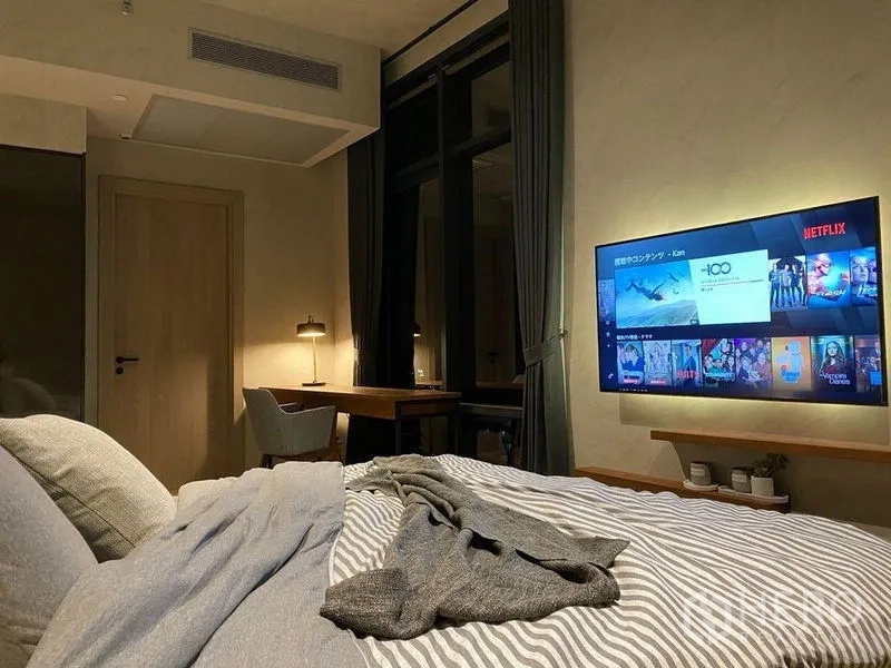 Condominium for rent or sale in Watthana, Bangkok - Cozy bedroom with bed, study desk, and wall-mounted TV, framed by dark curtains and soft lighting.