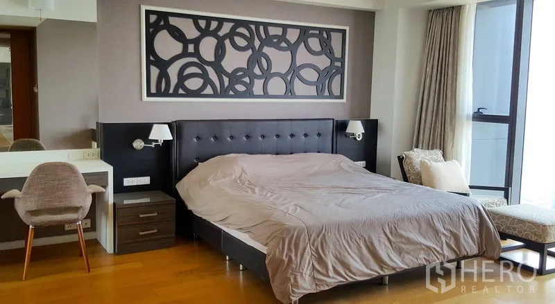 Condominium for rent in Sathon, Bangkok - Master bedroom with upholstered headboard, bedside lamps, wall art, desk and chair, and a large window with drapes.