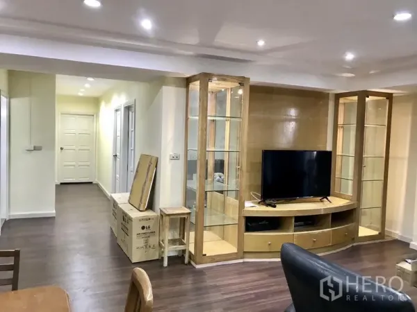 Condominium for rent in Watthana, Bangkok - Living room with glass display cabinets, TV console, hardwood-style floors, and hallway view.