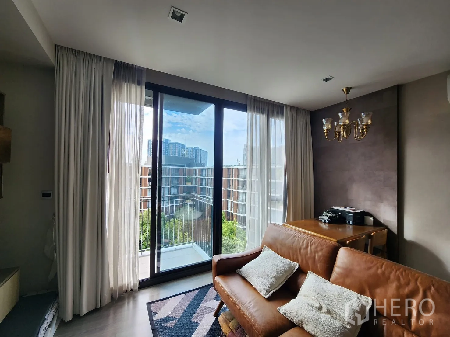 Condominium for rent in Watthana, Bangkok - Sofa area beside wide balcony doors with sheer curtains and chandelier, overlooking the city.