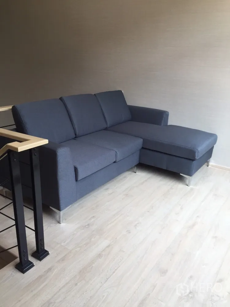 Townhouse for rent in Suan Luang, Bangkok - Bright living area with a gray L-shaped sofa and light wood floors beside the staircase.