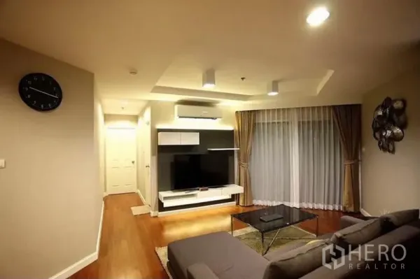 Condominium for rent in Huai Khwang, Bangkok - Cozy lounge with TV wall unit, recessed ceiling lights and full-height curtains leading to a balcony.