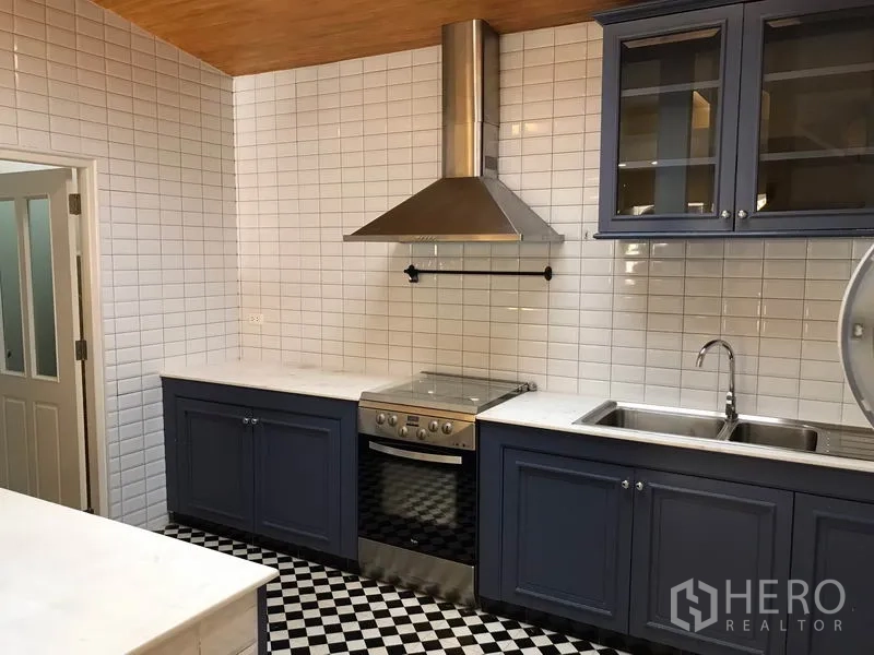 Townhouse for rent in Wang Thonglang, Bangkok - Fitted kitchen with navy cabinetry, subway-tile walls, marble countertops, stainless stove and hood, and checkerboard floor.