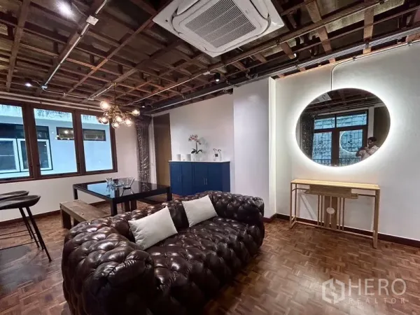 Apartment for rent in Khlong Toei, Bangkok - Chic living room with tufted leather sofa, glass dining table, exposed ceiling beams and a backlit round mirror.