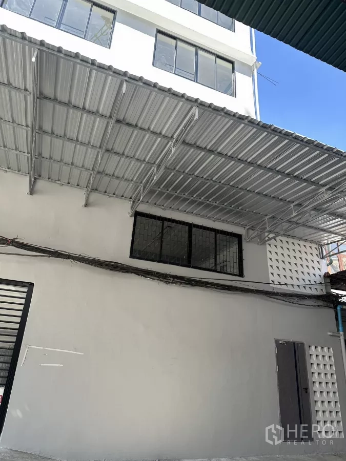 办公空间 出租在Phra Khanong, Bangkok - 低层建筑外立面，金属雨棚与黑框窗，蓝天下一角。