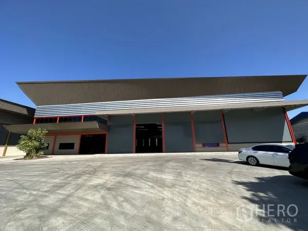 Factory for rent in Chachoengsao - Modern factory facade with high gabled roof, multiple roller shutter bays and a wide concrete yard under a clear sky in Bang Pakong.