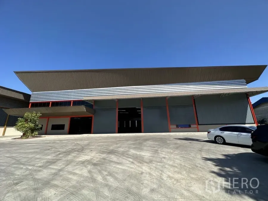 Factory for rent in Chachoengsao - Modern factory facade with high gabled roof, multiple roller shutter bays and a wide concrete yard under a clear sky in Bang Pakong.