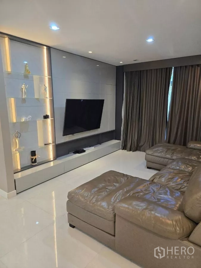 Detached House for rent or sale in Bang Phli, Samut Prakan - Cozy living room featuring a sectional sofa, wall-mounted TV, and illuminated display shelves.
