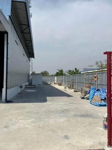 Warehouse for rent in Nonthaburi - Concrete driveway beside a metal-clad warehouse with secure fence and open loading yard in Sai Noi, Nonthaburi.